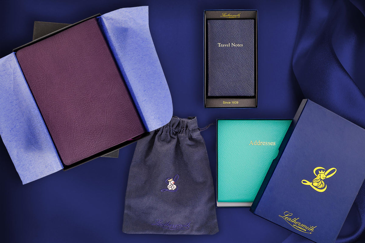 Leathersmith gift selection featuring a purple leather notebook, navy Travel Notes book and turquoise address book, with branded gift boxes and a drawstring pouch on a navy background.